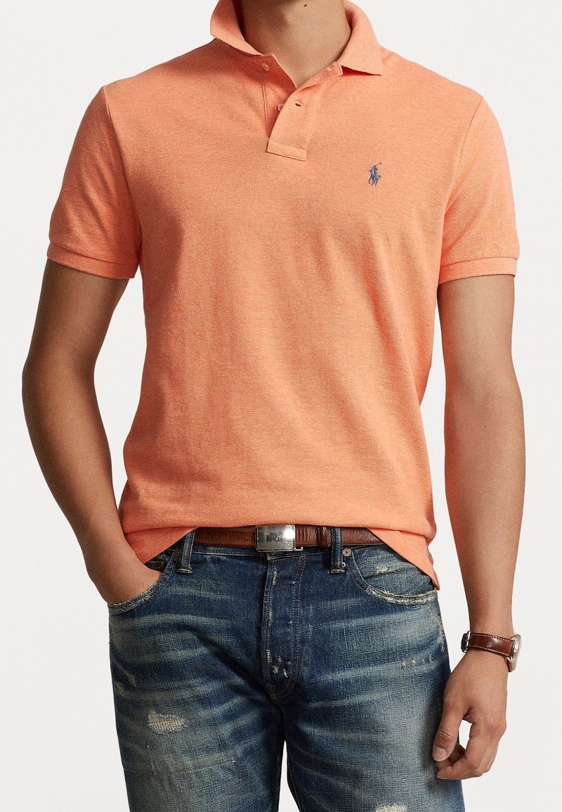 Homme portant un polo couleur pêche à manches courtes, un jean bleu déchiré, une ceinture marron et une montre-bracelet marron, debout avec une main dans la poche.