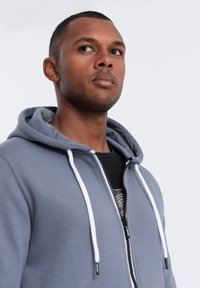 Hoodie zippé gris avec cordons blancs, affichant une texture lisse et une poche kangourou. Porté par-dessus un t-shirt noir à motifs.