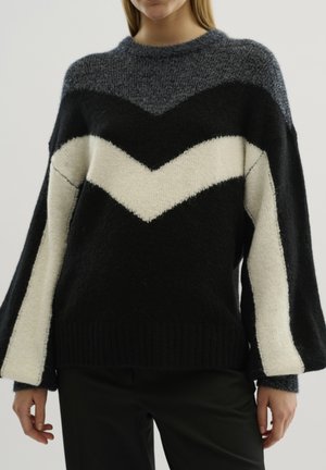 Pull en tricot à motif chevron noir et blanc, avec un col rond, des manches longues évasées et une finition douce et texturée.
