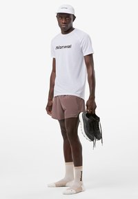 Weißes T-Shirt mit kurzen Ärmeln und schwarzem Logo, hellbraune Shorts, beige und weiße Socken, hält schwarze Sportschuhe; minimalistisches Design.