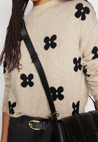 Beiger Strickpullover mit schwarzen Blumenmustern, rundem Ausschnitt und langen Ärmeln. Schwarze Schultertasche mit gewebtem Design und goldfarbenen Beschlägen.