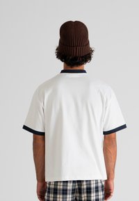 Man wearing a brown knit beanie, white short-sleeve shirt with black trim, and black-and-white plaid pants, facing away against a plain background.