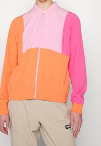 Veste colorée composée de panneaux orange, rose et rose clair. Le matériau a une texture lisse avec une taille et des poignets élastiques. Fermeture éclair.