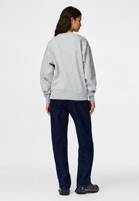 Moteris stovinti nugara, vilkinti šviesiai pilką džemperį, tamsiai mėlynas džinsas ir juodus sportinius batelius ant paprasto balto fono.