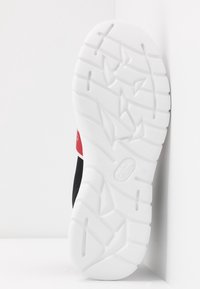 Semelle blanche texturée d'une basket avec des rainures profondes et un petit logo en relief, inclinée verticalement contre un fond blanc en coin.