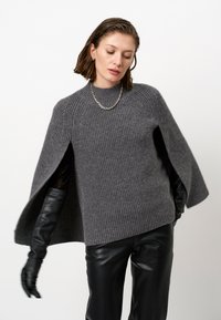 Grijze gebreide trui met een hoge hals en brede kaapsleeves, gecombineerd met zwarte leren broek en handschoenen. Geweven stofdetail zichtbaar.