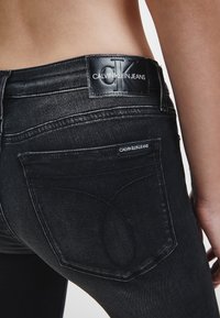 Jeans de mezclilla negra con una textura suave, un parche con el logo de Calvin Klein en la cintura y detalles de costura sutiles en los bolsillos.