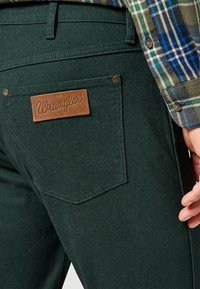 Donkergroene Wrangler jeans met een leren logo label, koperen klinknagels en een achterzak met een geborduurd detail. Getextureerde denim stof.