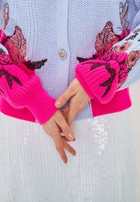 Cardigan lavorato a maglia con corpo azzurro chiaro, polsini a costine rosa con ricami floreali e dettaglio con bottoni argentati. Sotto, una gonna bianca con paillettes.