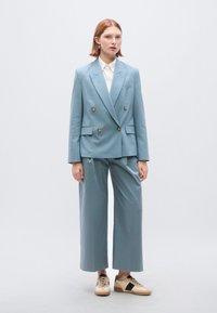 Lichtblauw dubbelgerijpt colbert met decoratieve knopen, gecombineerd met bijpassende broek met wijde pijpen en beige sneakers met zwarte accenten.