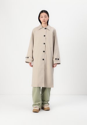 Cappotto lungo beige in tessuto con bottoni neri, colletto classico, maniche larghe e tasche laterali, abbinato a pantaloni verdi e calzature beige.