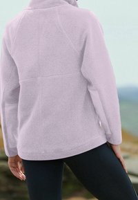 Chaqueta de forro polar lavanda claro con una superficie texturizada, bolsillos laterales con cremallera y un cuello alzado. Presenta mangas ranglan y un corte relajado.