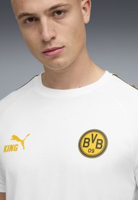 Unge mand if�ørt en hvid Borussia Dortmund fodboldtrøje med Puma-logo og teksten "KING", mod en neutral grå baggrund.