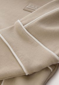Tissu beige avec des détails en passepoil blanc et un petit patch rectangulaire avec des lettres en relief partiellement visibles en arrière-plan.
