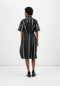 Rochie neagră, cu dungi verticale albe, cu mâneci scurte, talie accentuată printr-o curea și lungime până la jumătatea gambei. Fabricată dintr-un material ușor, cu o textură netedă.
