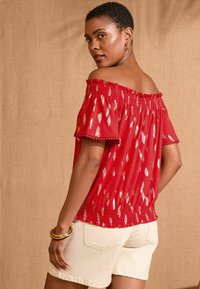 Raudonas off-the-shoulder top'as su sidabriniais raštais, susmulkinta detalėmis ir volanų kraštais. Derinamas su šviesiais beige šortais ir apvaliais auksiniais apyrankėmis.