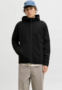 Veste noire à capuche conçue avec une fermeture éclair à l'avant, équipée de poches latérales et d'un tissu doux et lisse. Portée sur des vêtements beiges avec une casquette bleue.