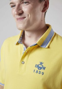 Polo shirt jaune en coton, avec un logo bleu brodé, deux boutons au col et un détail de col intérieur à motif.