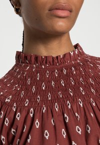 Burgundy smocked blouse with a high ruffled collar, adorned with white geometric patterns and a textured fabric finish.