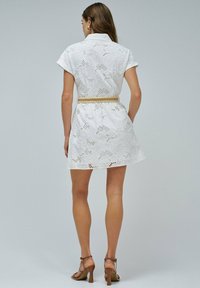 Mujer de pie de espaldas a la cámara, vestida con un vestido blanco de encaje de manga corta y un cinturón beige, acompañado de sandalias marrones de tacón.