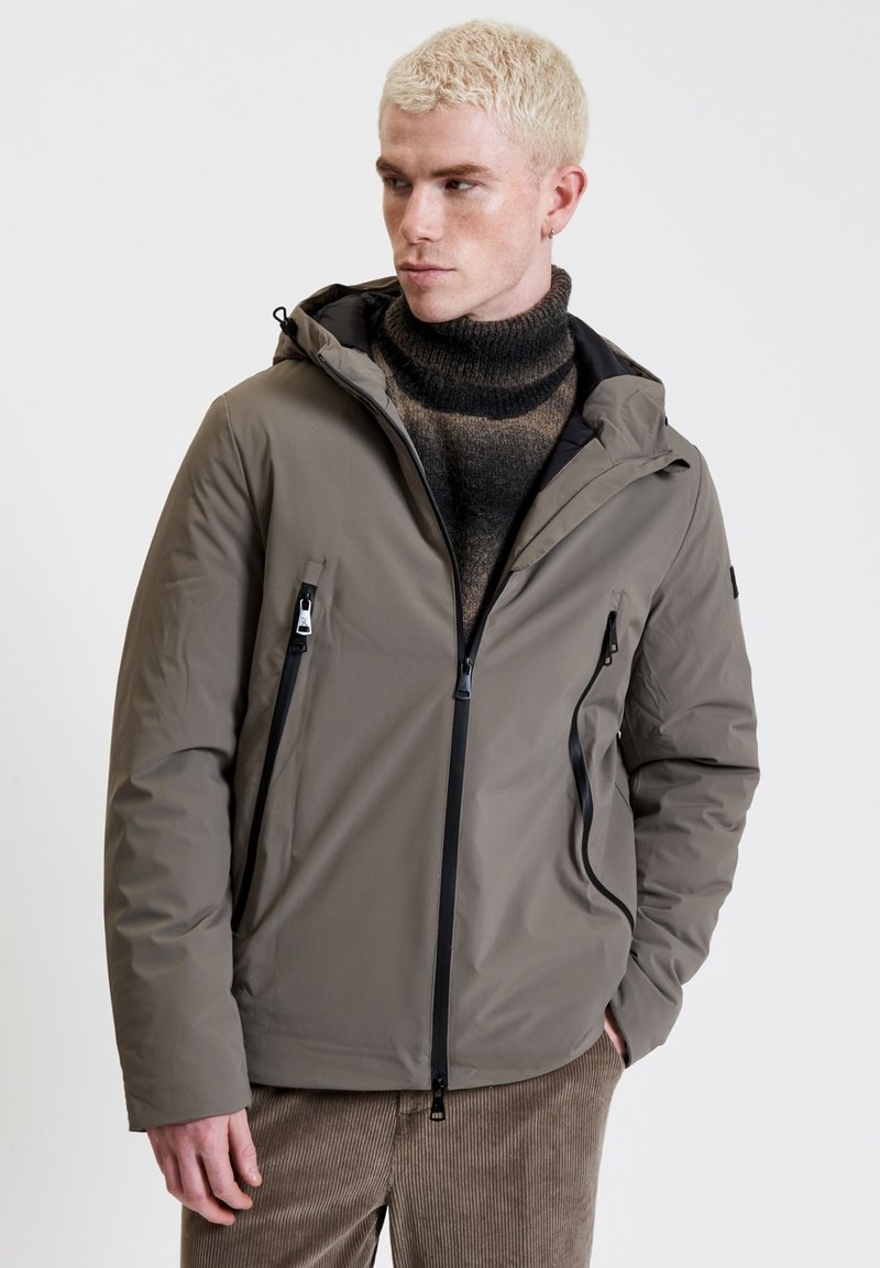 Giacca khaki con cappuccio e tasche con zip, realizzata in materiale liscio e impermeabile. Caratterizzata da un colletto alto e dettagli neri sulle zip.