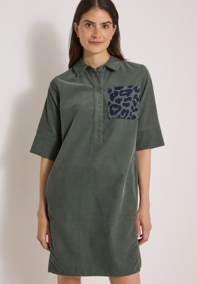 Jeune femme portant une robe ample en velours côtelé vert foncé avec des manches courtes et une poche poitrine à motif, debout devant un mur clair uni.