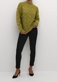 Olivgrüner gestrickter Pullover mit wellenförmigem Muster, hohem Kragen und langen Ärmeln; kombiniert mit schwarzen Slim-Fit-Jeans und schwarzen Pumps.