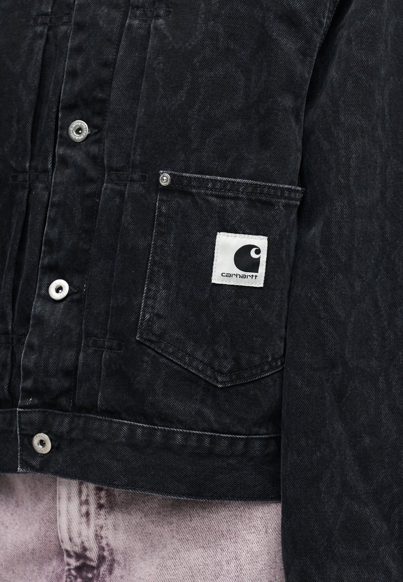 Giacca di denim nera con tasca frontale che presenta una toppa con il logo Carhartt bianco, indossata sopra jeans sbiaditi di colore viola chiaro.