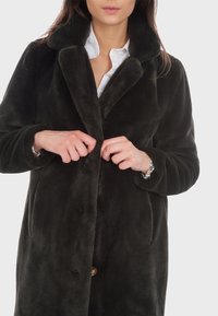 Manteau en fourrure noire avec une texture douce, col à revers, poches avant et fermeture à bouton doré. Le modèle boutonne le manteau.