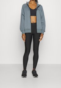 Grå zip-up idrottsjacka, svart sportbh, svarta leggings och svarta sneakers. Jackan har en hög krage och ribbade manschetter.