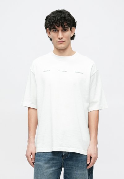 White cotton t-shirt with short sleeves, round neck, featuring printed text on the front. Worn with faded blue denim jeans.