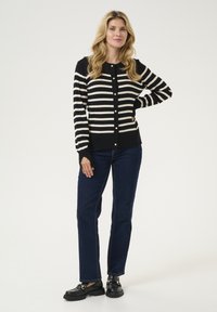 Juodai baltas dryžuotas kardiganas su mygtukų užsegimu, juodomis rankovėmis ir kišenėmis, derinamas su tamsiai mėlynomis džinsomis ir juodais batais. Lygi tekstūra.