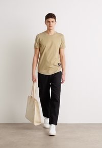 Tricou din bumbac bej cu mâneci scurte, asortat cu pantaloni negri. Modelul poartă o geantă tote bej și adidași albi.
