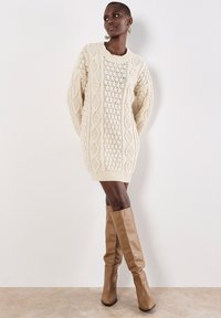 Flauschiges, grob gestricktes Sweaterkleid in Creme mit langen Ärmeln und strukturierten Mustern. Kombiniert mit hohen, sandfarbenen Lederstiefeln mit Absatz.