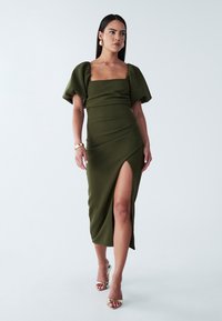 Maslinasto zeleni haljina s napuhanim rukavima, prilagođenim korzetom i bočnim prorezom. Izrađena od glatkog materijala s elegantnim dizajnom i minimalnom teksturom.