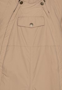Beige isolierte Latzhose mit verstellbaren Schulterträgern, einer Fronttasche und Metall-Druckknöpfen; glattes, wasserabweisendes Material.