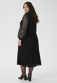 Juoda maxi suknelė su ilgomis nėrinių rankovėmis, suplota liemenėle, sluoksniuota sijonu ir derinama su juodais bateliais ant kulno. Lygi tekstūra, elegantiškas dizainas.