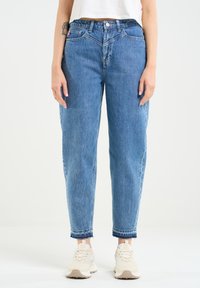 Blaue Denim-Jeans mit lockerer Passform, hoher Taille, klassischem Fünf-Taschen-Design und ausgefranstem Saum. Getragen mit hellen Sneakern.