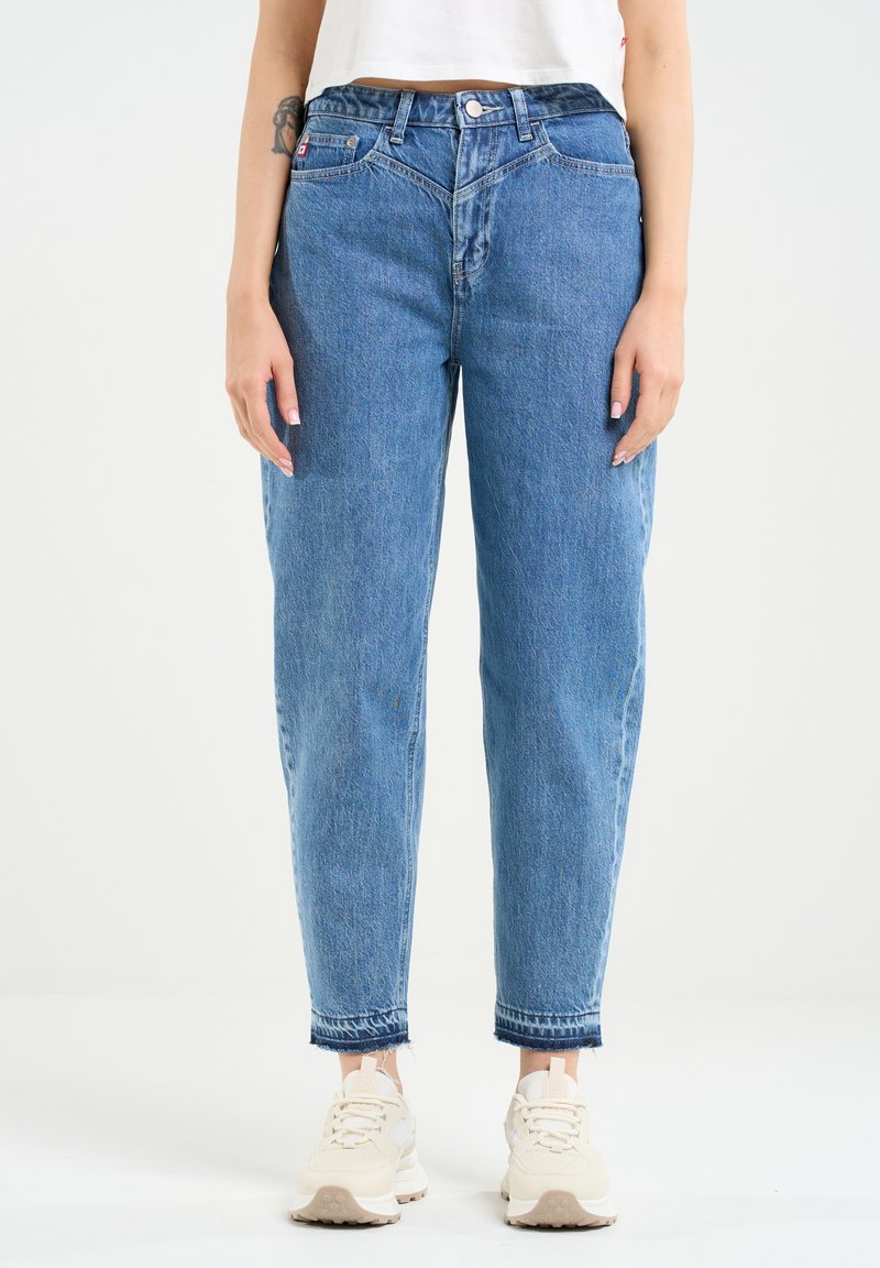 Blaue Denim-Jeans mit lockerer Passform, hoher Taille, klassischem Fünf-Taschen-Design und ausgefranstem Saum. Getragen mit hellen Sneakern.