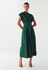 Žalia, iki kelių siekianti suknelė su trumpomis rankovėmis, plisuota konstrukcija, aukšta apykakle ir prigludusia juosmena; derinama su smailiais, vytinėmis tekstūromis papuoštais bateliais.