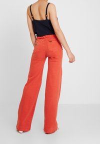 Pantaloni in denim arancione a gamba larga e alta vita, con un'etichetta del marchio visibile sulla tasca posteriore. Materiale in cotone con una texture liscia.