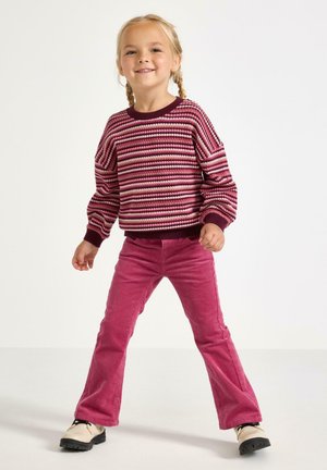 Gebreide trui in bordeauxrood, roze en crèmekleurige strepen; geribbelde manchetten. Strakke roze ribfluwelen broek en crèmekleurige schoenen met zwarte zolen.