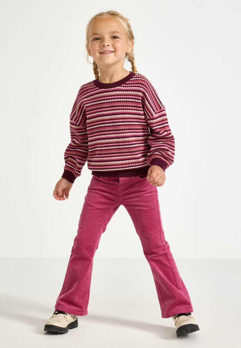 Strikket sweater i bordeaux, pink og creme striber; ribbede manchetter. Tætsiddende pink corduroy bukser og creme sko med sorte såler.