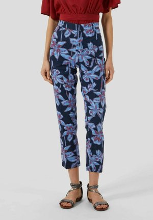 Nainen pukeutuneena sinisiin kukkakuosillisiin lyhytlahkeisiin housuihin, punaiseen lyhythihaiseen topiin ja mustiin nilkkaremmi-sandaaleihin yksivärisellä taustalla.