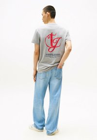 Camiseta de algodón gris con un gran logotipo rojo en la parte trasera, combinada con jeans de pierna ancha azul claro y zapatillas blancas.