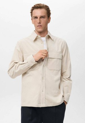 Camisa de pana beige con collar, cierre de botones, dos bolsillos en el pecho y mangas largas. Tejido texturizado con rayas verticales.