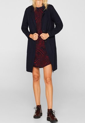 Kobieta w czarnym płaszczu do kolan nałożonym na czerwono-czarną sukienkę we wzory, w bordowych sznurowanych botkach, stojąca na tle jednolitego szarego tła.