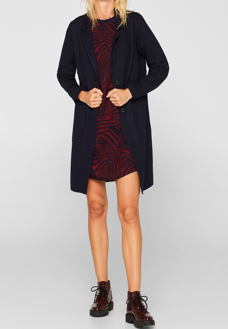 Femme portant un manteau noir arrivant aux genoux, par-dessus une robe rouge et noire à motifs, avec des bottines bordeaux à lacets, debout devant un fond gris uni.