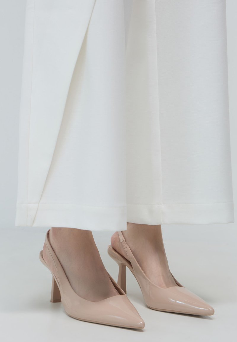 Pieds chaussés de talons hauts en cuir verni nude avec une bride derrière, associés à un pantalon large blanc s'arrêtant au-dessus des chevilles.