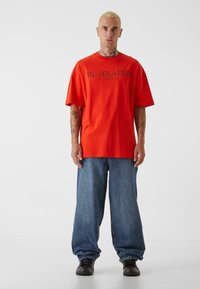 Rød, oversized, bomulls-T-skjorte med svart tekst "REVELATION 12:07:12" på forsiden. Bært med baggy, blå jeans og svarte sko.